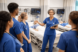 Nurse educator teaching a group of nursing students in a clinical simulation lab with patient mannequins and monitoring equipment.