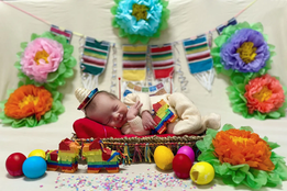 A newborn in the NICU dressed in a tiny Fiesta-themed outfit sleeps on a colorful blanket surrounded by festive decorations, including paper flowers, mini piñatas and bright eggs