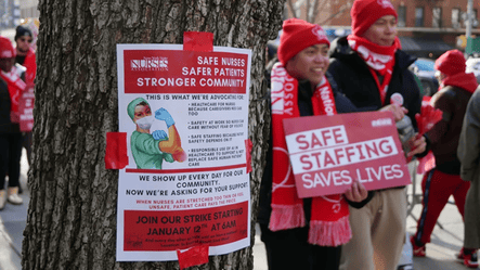 Nurses wearing red strike apparel hold signs reading “Safe Staffing Saves Lives” during a nurse strike, with a strike flyer taped to a tree in the foreground.