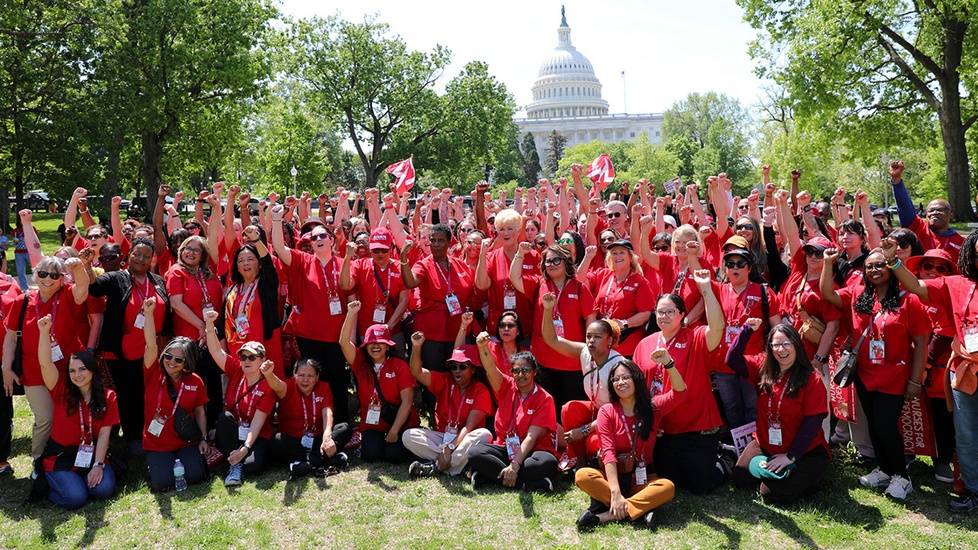 A large group of nurses wearing red shirts raises their fists in solidarity in front of the U.S. Capitol. They are gathered on the lawn, demonstrating support for safe staffing legislation.