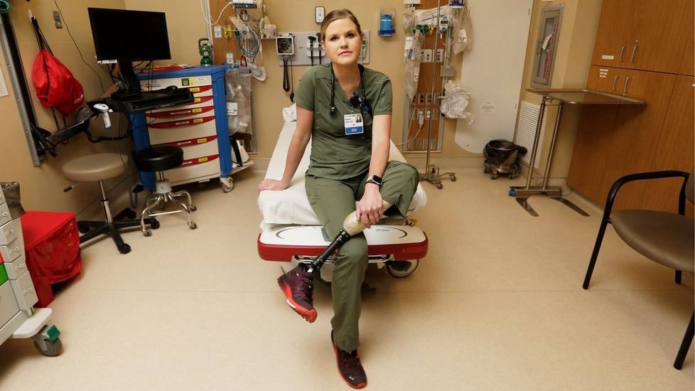 ER nurse Hannah Cvancara sitting on a hospital stretcher in an emergency room, wearing green scrubs and showing her prosthetic left leg.