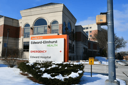 Front entrance of Edward Hospital in Naperville, Illinois, with a large Edward-Elmhurst Health sign and emergency directions, surrounded by snow-covered ground and bushes on a clear winter day.