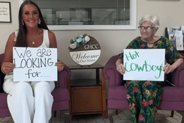 A nurse and an older woman sit holding signs that read “We are looking for…” and “Hot Cowboys” at Gainesville Health Care Center.