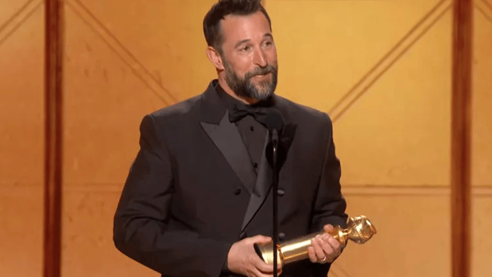 Noah Wyle in a dark tuxedo standing at a microphone at the Golden Globes awards stage, holding a statuette in both hands.