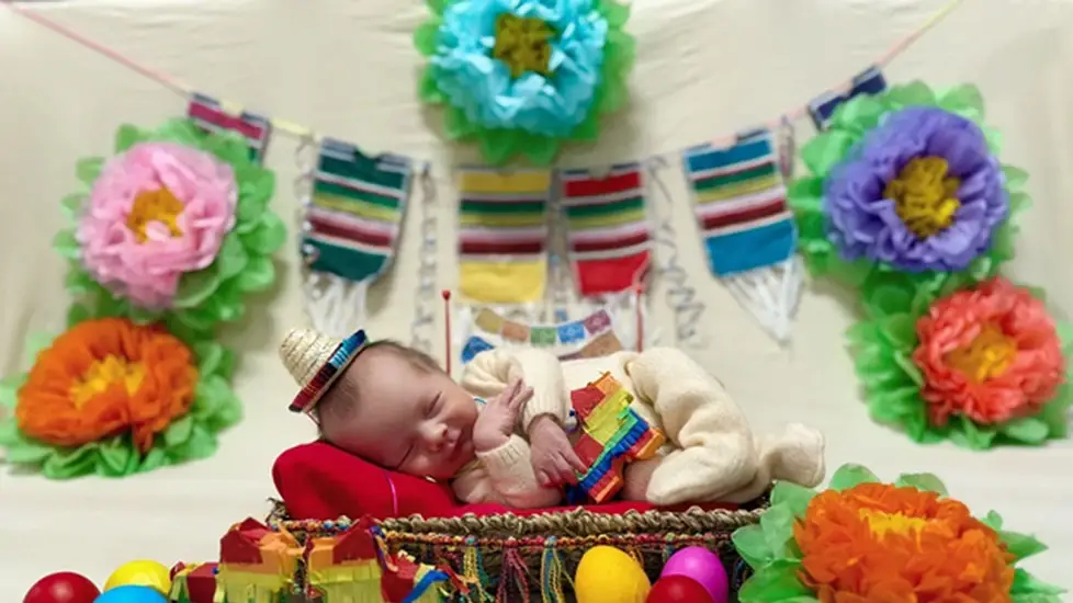 A newborn in the NICU dressed in a tiny Fiesta-themed outfit sleeps on a colorful blanket surrounded by festive decorations, including paper flowers, mini piñatas and bright eggs