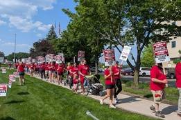 A large group of nurses and supporters march along a sidewalk outside a hospital during a sunny day protest. Many wear red shirts with union slogans and carry signs reading “PATIENTS BEFORE PROFITS.”