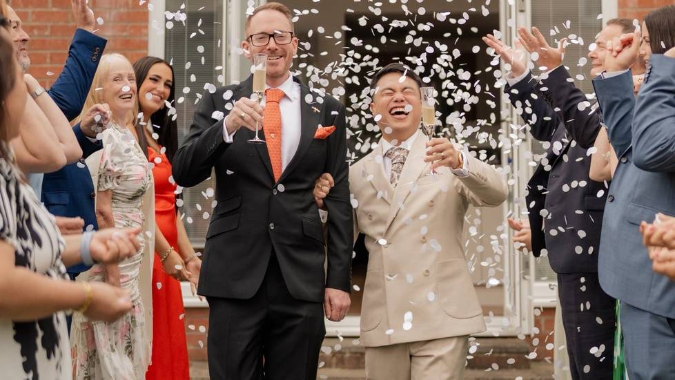 Nurse Even and his husband Paul Szom walk hand in hand while smiling and holding champagne glasses as friends and family shower them with white confetti outside their wedding venue.