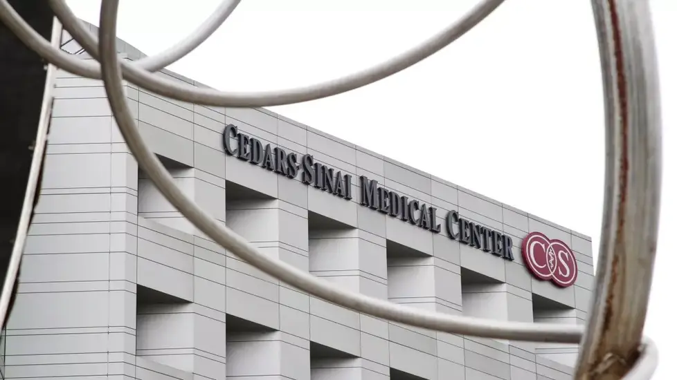 The exterior of Cedars-Sinai Medical Center in Los Angeles, showing the hospital's name and logo on a modern, white building, partially framed by curved metal sculpture elements in the foreground