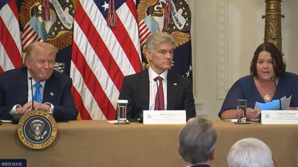 Donald Trump, Dr. Oz and Amy Gleason sit at a table with microphones and nameplates, speaking at a White House event. Behind them are multiple American flags and presidential seals.