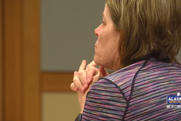 A woman, identified as Alaska nurse practitioner Kris Kile, sits in a courtroom wearing a striped top, her hands clasped near her face in a contemplative pose. The image is sourced from Alaska’s News