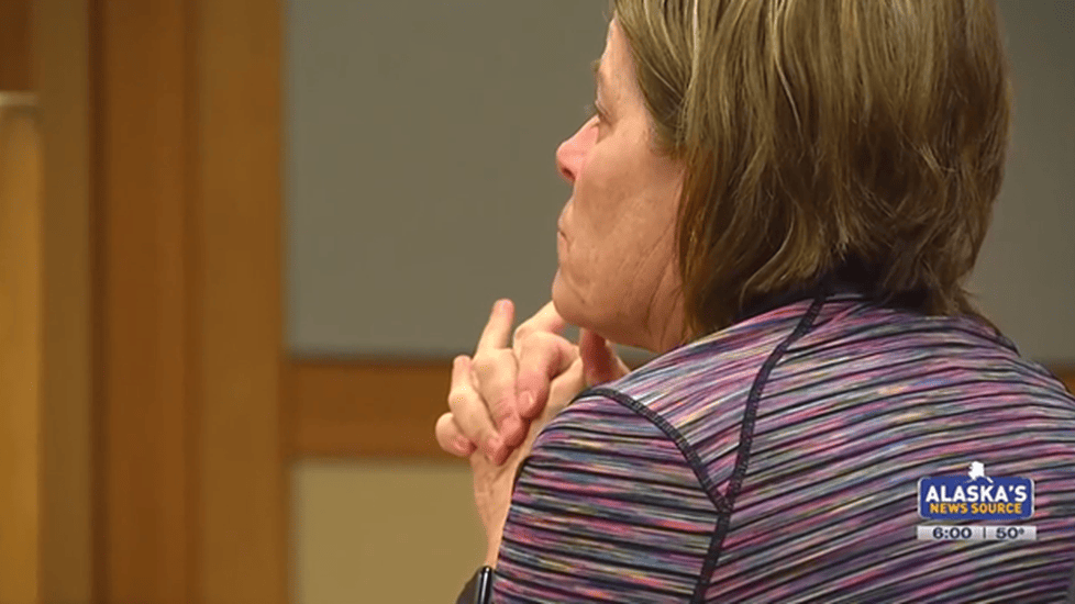A woman, identified as Alaska nurse practitioner Kris Kile, sits in a courtroom wearing a striped top, her hands clasped near her face in a contemplative pose. The image is sourced from Alaska’s News