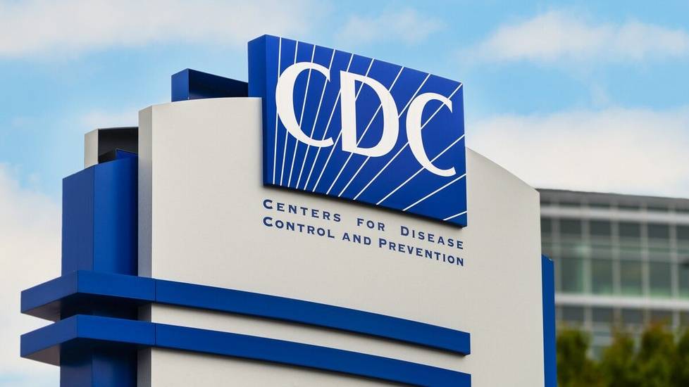 CDC (Centers for Disease Control and Prevention) sign outside headquarters, with blue and white logo against a partly cloudy sky.