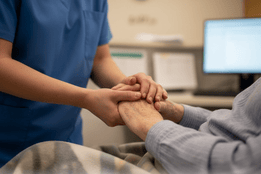 A nurse in blue scrubs gently holds the hand of an elderly patient, conveying compassion in a long-term care setting. Their faces are not visible.