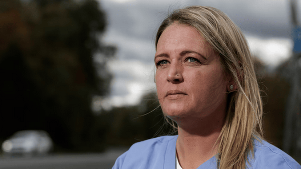 Andrea Elia wearing light blue medical scrubs sits outdoors near a roadside, looking thoughtful into the distance. The sky is cloudy, and blurred trees and a car appear in the background.