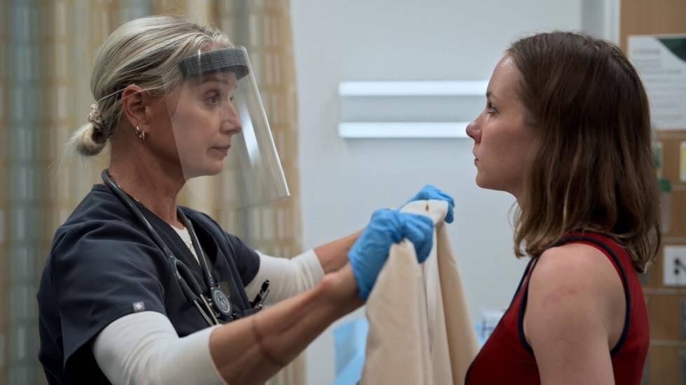 The Pitt's Sexual Assault Nurse Examiner Dana Evans wearing protective gear prepares evidence collection materials while speaking with a patient in an exam room.