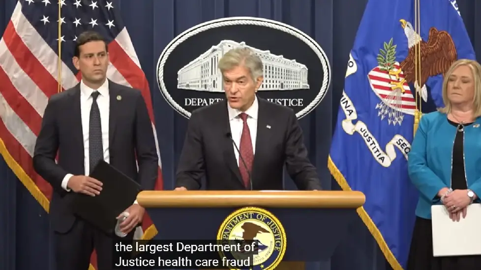 U.S. Department of Justice officials stand at a podium during a press conference announcing the largest healthcare fraud takedown in DOJ history, with American and DOJ flags in the background.