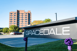 A large, multi-story brick building stands in the background, with a parking lot and several cars in front. In the foreground, a black sign with bold white letters reads "GRACEDALE"