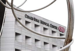 The exterior of Cedars-Sinai Medical Center in Los Angeles, showing the hospital's name and logo on a modern, white building, partially framed by curved metal sculpture elements in the foreground