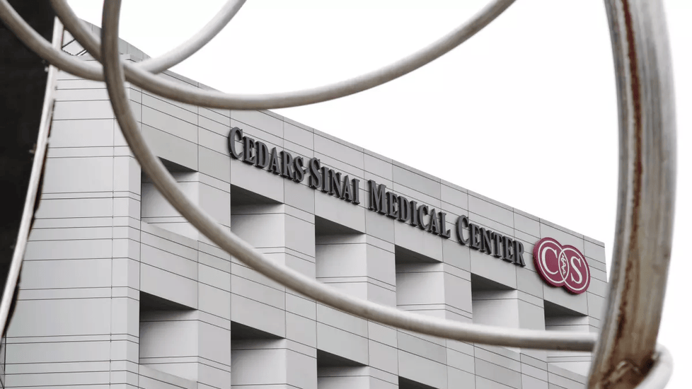 The exterior of Cedars-Sinai Medical Center in Los Angeles, showing the hospital's name and logo on a modern, white building, partially framed by curved metal sculpture elements in the foreground