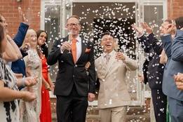 Nurse Even and his husband Paul Szom walk hand in hand while smiling and holding champagne glasses as friends and family shower them with white confetti outside their wedding venue.