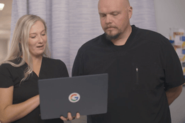 Two nurses in black scrubs review patient information together on a Google-branded laptop in a hospital hallway, illustrating collaboration with technology in healthcare.