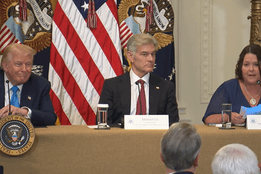 Donald Trump, Dr. Oz and Amy Gleason sit at a table with microphones and nameplates, speaking at a White House event. Behind them are multiple American flags and presidential seals.