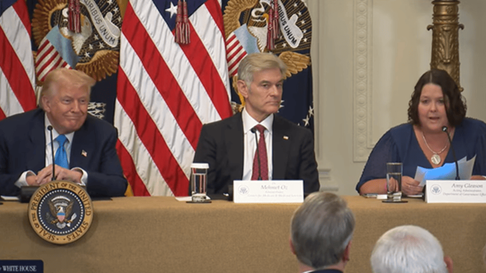 Donald Trump, Dr. Oz and Amy Gleason sit at a table with microphones and nameplates, speaking at a White House event. Behind them are multiple American flags and presidential seals.