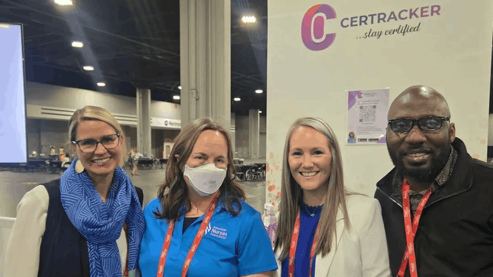 CerTracker team smiling at conference booth: two women in lanyards (one blue shirt, one white blazer), man in glasses and navy jacket. Purple logo backdrop.