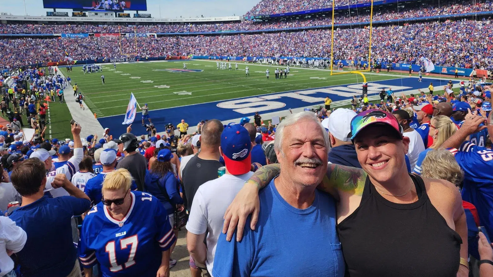 Brian Dodge smiling at a Buffalo Bills game with the stadium crowd behind them.