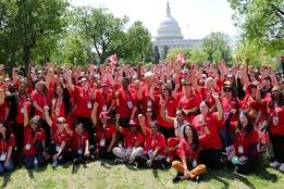 A large group of nurses wearing red shirts raises their fists in solidarity in front of the U.S. Capitol. They are gathered on the lawn, demonstrating support for safe staffing legislation.