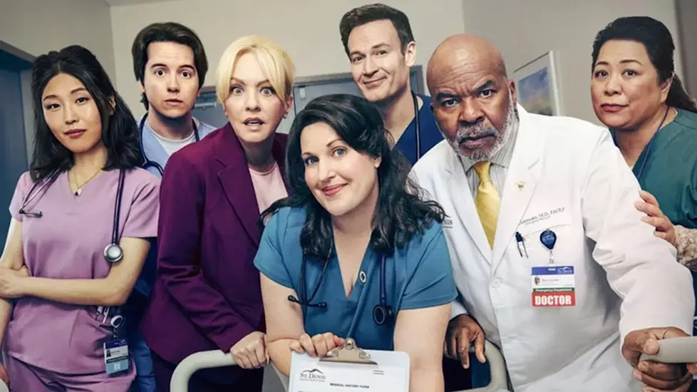 The cast of St. Denis Medical poses in a hospital hallway, dressed in scrubs and lab coats, capturing the show’s comedic hospital vibe.