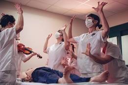 Nurses in white scrubs perform expressive dance movements around a patient on a hospital bed during Tara Rynders' "First Do No Harm."