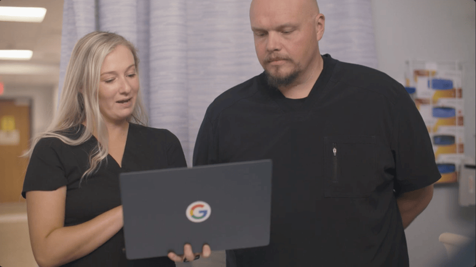Two nurses in black scrubs review patient information together on a Google-branded laptop in a hospital hallway, illustrating collaboration with technology in healthcare.
