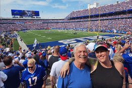 Brian Dodge smiling at a Buffalo Bills game with the stadium crowd behind them.